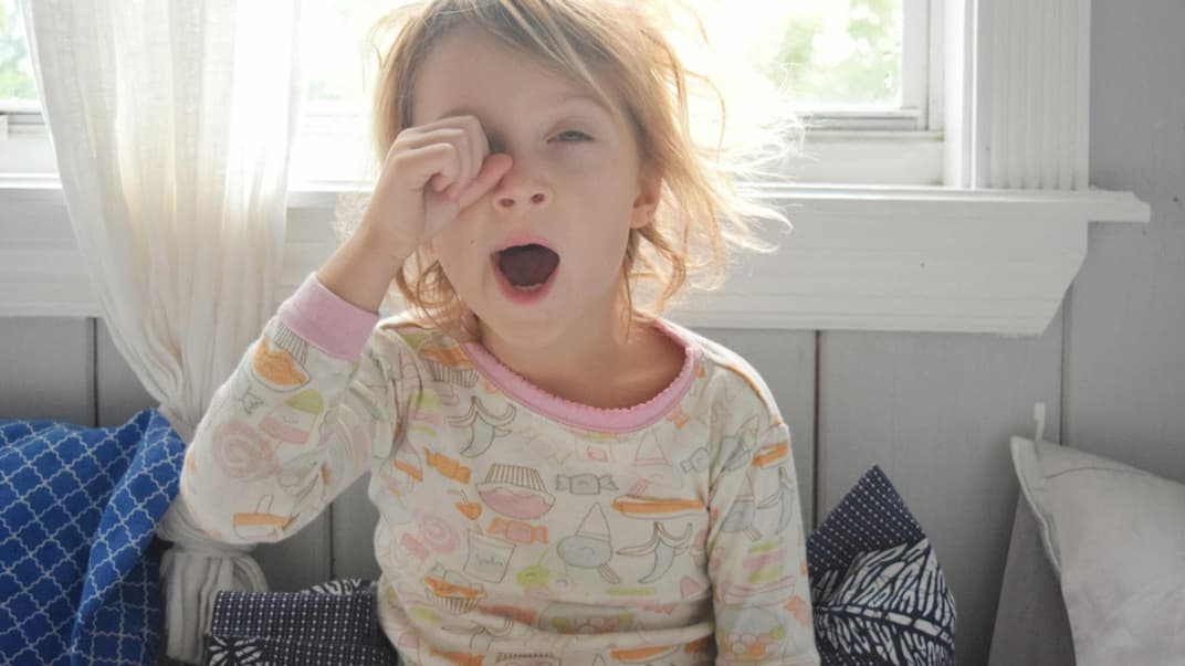 Gerade für Kinder ist die Zeitumstellung nicht immer leicht. Blondes Mädchen reibt sich am Morgen die Augen und gähnt