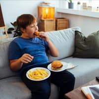 Fast-Food sollte nicht allzu häufig auf dem Speiseplan für Kinder stehen. Dicker Junge isst Burger und Pommes auf dem Sofa.