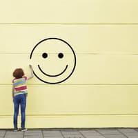 Kinder sind von Natur aus glücklich - und dieses Glück gilt es zu stärken. Mädchen malt Smiley an gelbe Wand.