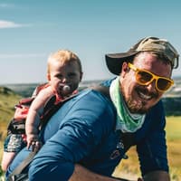 Auf einem Nordic-Walking-Familienausflug kann man sich mit dem Tragen des Babys abwechseln. Fröhlicher Vater trägt Baby auf dem Rücken durch die Landschaft.