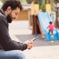 Neulich auf dem Spielplatz: Väter, die nebenbei arbeiten, erhalten Anerkennung. Mütter dagegen Missbilligung. Vater mit Handy auf Spielplatz.