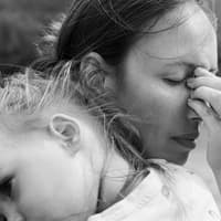 Viele Mütter stehen im Alltag unter Dauerbelastung. Eine verzweifelte Mutter hält ihre Tochter auf dem Arm, während sie die Stirn runzelt, die Augen schließt und sich an die Nase fässt.