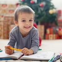 Weihnachtliche Ausmalbilder sind bei Kindern sehr beliebt. Ein kleiner Junge liegt vor dem Weihnachtsbaum auf dem Boden und malt Bilder..