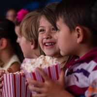 Der erste Ausflug ins Kino oder auch eingekuschelt auf dem Sofa – ein schöner Kinderfilm kann Klein und Groß Spaß machen. Zwei Kinder sitzen im Kino mit einer Tüte Popcorn in der Hand.