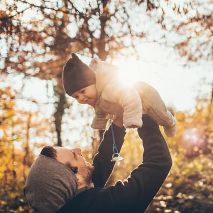 Was ist die richtige Babykleidung im Herbst? Papa hält Baby hoch im herbstlichen Wald