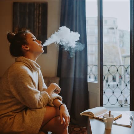 Frau sitzt auf einem Sofa am Fenster und raucht eine E-Zigarette. Ist E-Zigaretten-Dampf für Babys und Kinder gefährlich?
