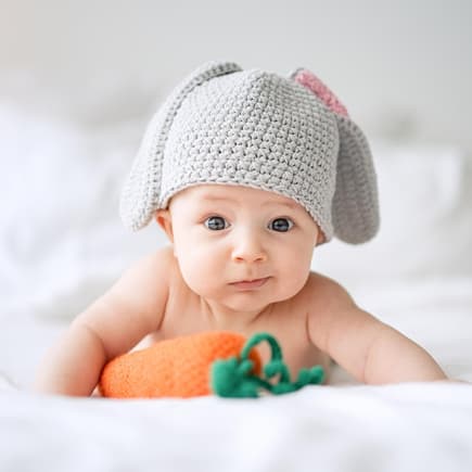 Osternamen für Babys können verschiedene Bedeutungen haben. Baby mit Hasenmütze vor Spielzeug-Möhre.