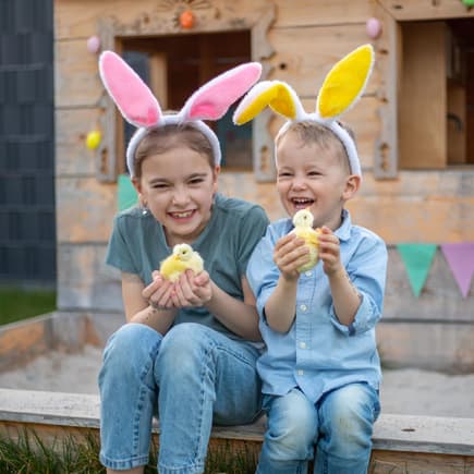 Ein Osterausflug macht gute Laune. Zwei Kinder mit Osterhasenohren und einem Küken in der Hand