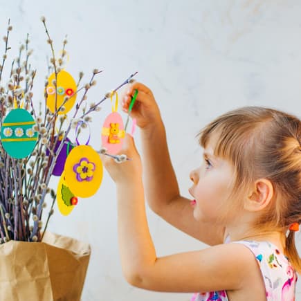 Ostereier lassen sich auf vielfältige Art und Weise basteln. Kleines Mädchen hängt selbst gebastelte Ostereier an den Strauch.