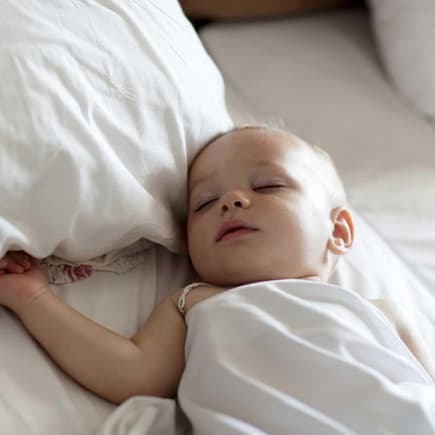 Fakt: Babys sollten noch nicht dauerhaft auf einem Kissen schlafen. Baby schläft in einem Bett mit großen Kissen.