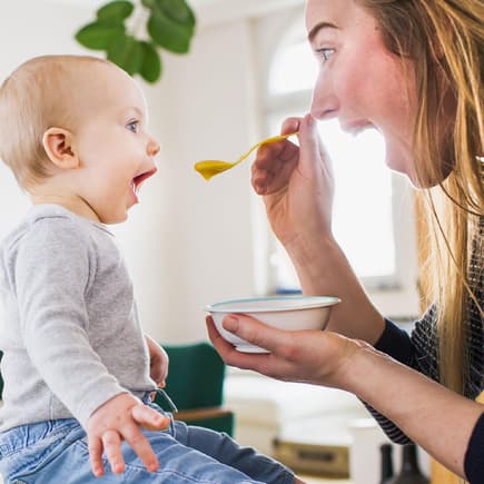 Der erste Brei sollte nicht völlig willkürlich gegeben werden. Hier kommt ein Beikostplan für Babyeltern. Mama füttert Baby mit Brei. Beide sperren den Mund auf.