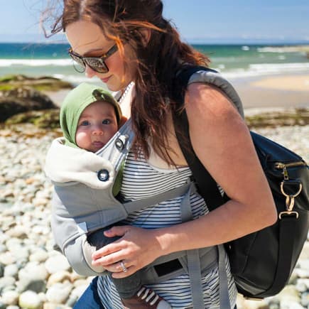 Im Sommer sollte euer Baby in der Trage weder schwitzen noch einen Sonnenbrand erleiden. Mutter mit Baby im Tragesitz am Strand