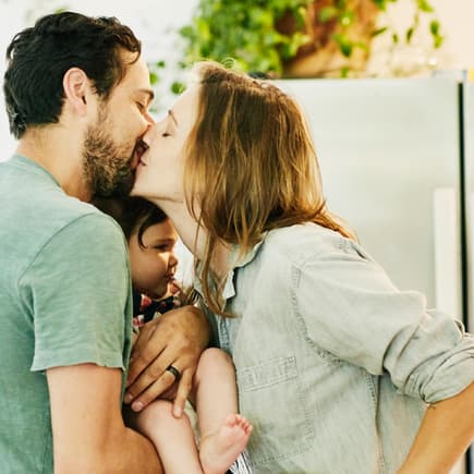 Sind die Eltern glücklich, profitieren davon auch die Kinder. Eltern küssen sich mit Kind auf dem Arm.