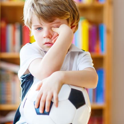 Bei Husten und Schnupfen liegt es im Ermessen der Eltern, ob sie ihr Kind in die Kita schicken oder lieber zu Hause lassen. Krankes Kind reibt sich die Augen mit Fußball im Arm.