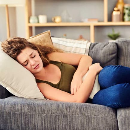 Regelbeschwerden – möglicherweise steckt eine Endometriose dahinter. Frau liegt mit Bauchschmerzen auf dem Sofa.