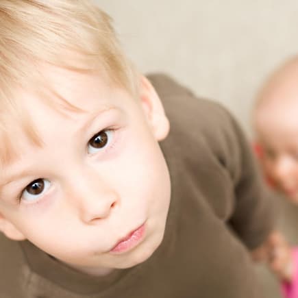 Wenn ein Kind ein Geschwisterchen bekommt, ist das anfangs nicht immer leicht. Ein kleiner Junge schaut genervt, während ein Baby sich an ihm hochzieht.
