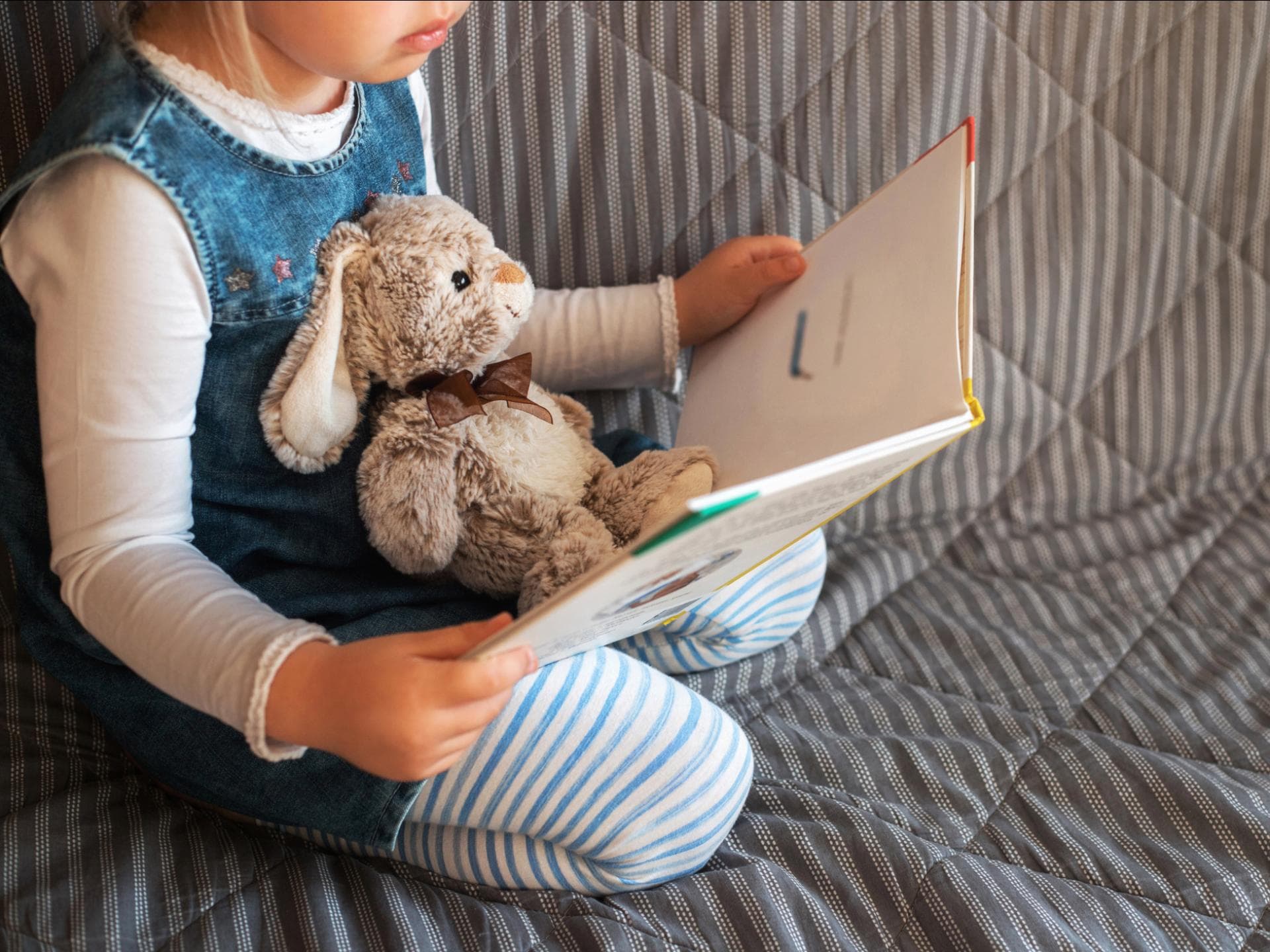 Die Bilder von Axel Scheffler erwecken viele Bücher zum Leben. Kleines Mädchen sitzt auf einem Sofa, ein Hasen-Kuscheltier auf ihrem Schoß, und hält ein aufgeschlagenes Buch in der Hand.