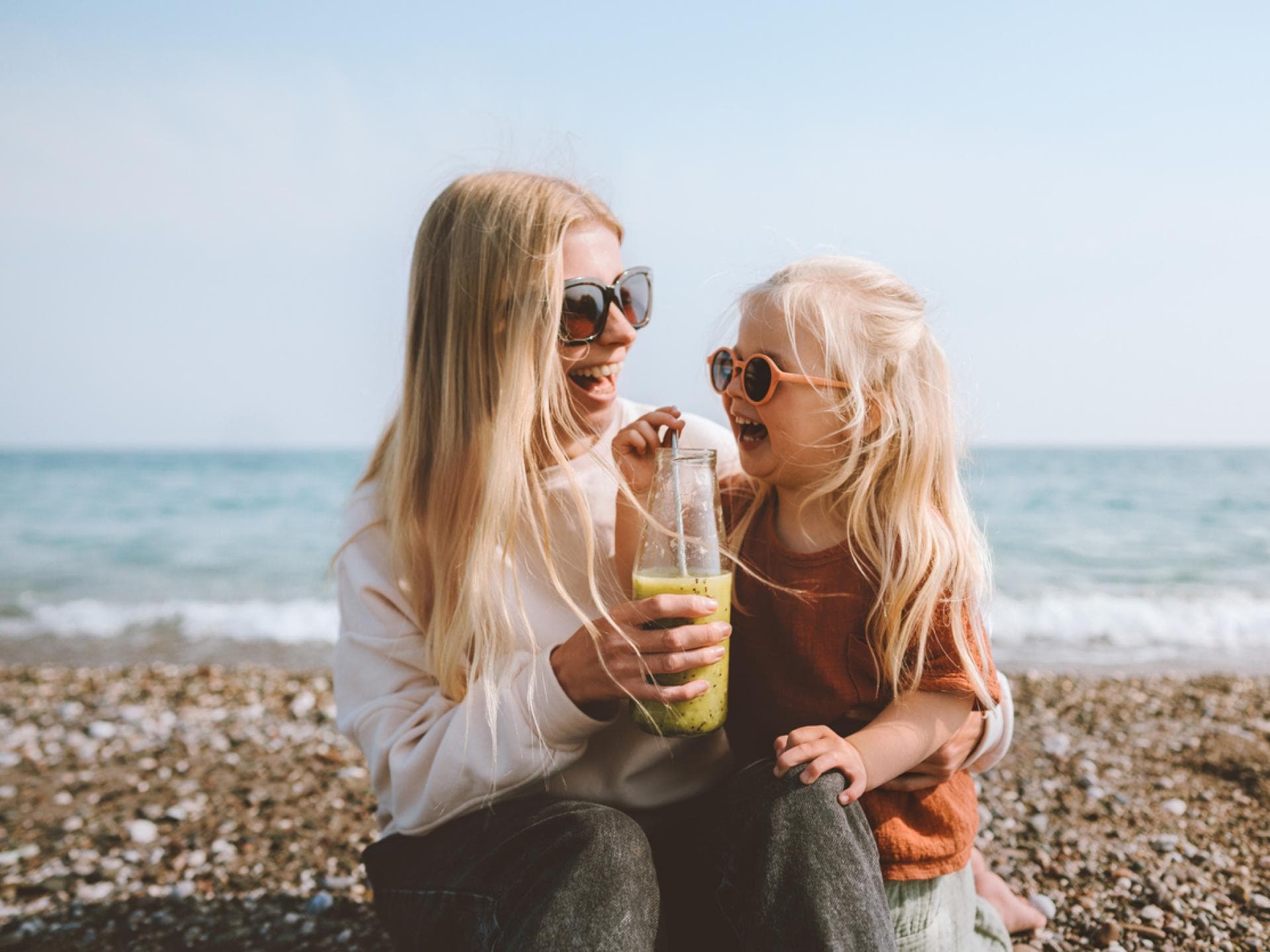 Mutter und Tochter sitzen fröhlich am schwedischen Strand, trinken einen Smoothie und lachen. Mutter und Tochter sitzen fröhlich am schwedischen Strand, trinken einen Smoothie und lachen.