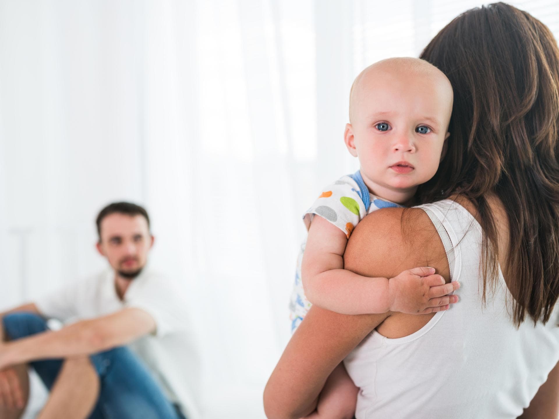 Wenn sich nur die Mutter ums Baby kümmert, ist Streit vorprogrammiert. Mutter hält Baby auf dem Arm, Vater sitzt im Hintergrund.