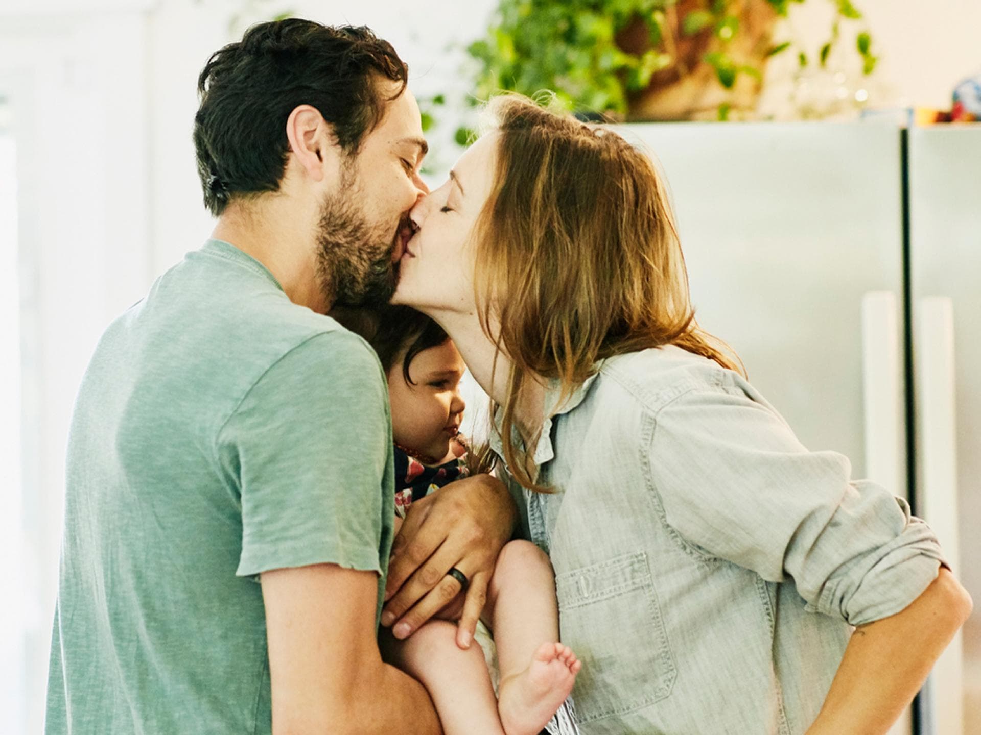 Sind die Eltern glücklich, profitieren davon auch die Kinder. Eltern küssen sich mit Kind auf dem Arm.