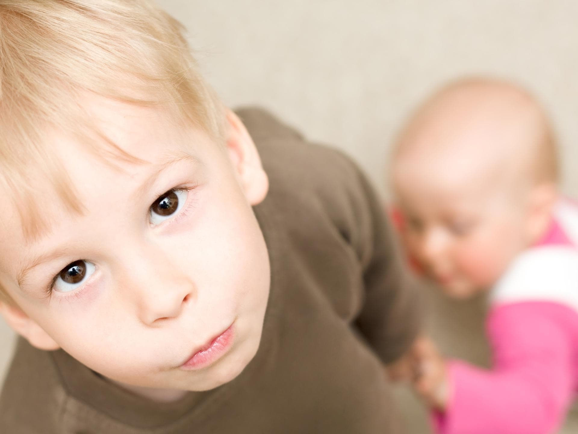 Wenn ein Kind ein Geschwisterchen bekommt, ist das anfangs nicht immer leicht. Ein kleiner Junge schaut genervt, während ein Baby sich an ihm hochzieht.